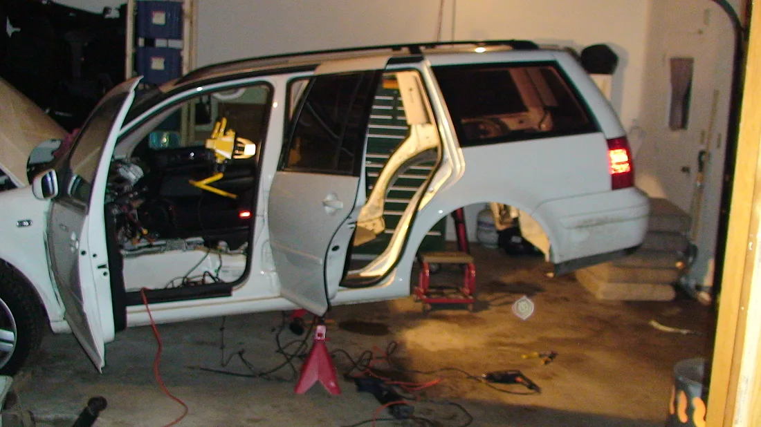 A Jetta wagon on jack stands, stripped to a bare shell with the rear floor cut out, garage floor visible through the car.